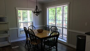 A dining room with a wooden table and chairs, a chandelier, and large windows with a view of trees.