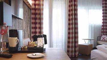 Quarto de hotel com uma mesa de madeira, cafeteira, chávena, bandeja com aperitivos, cama e cortinas.