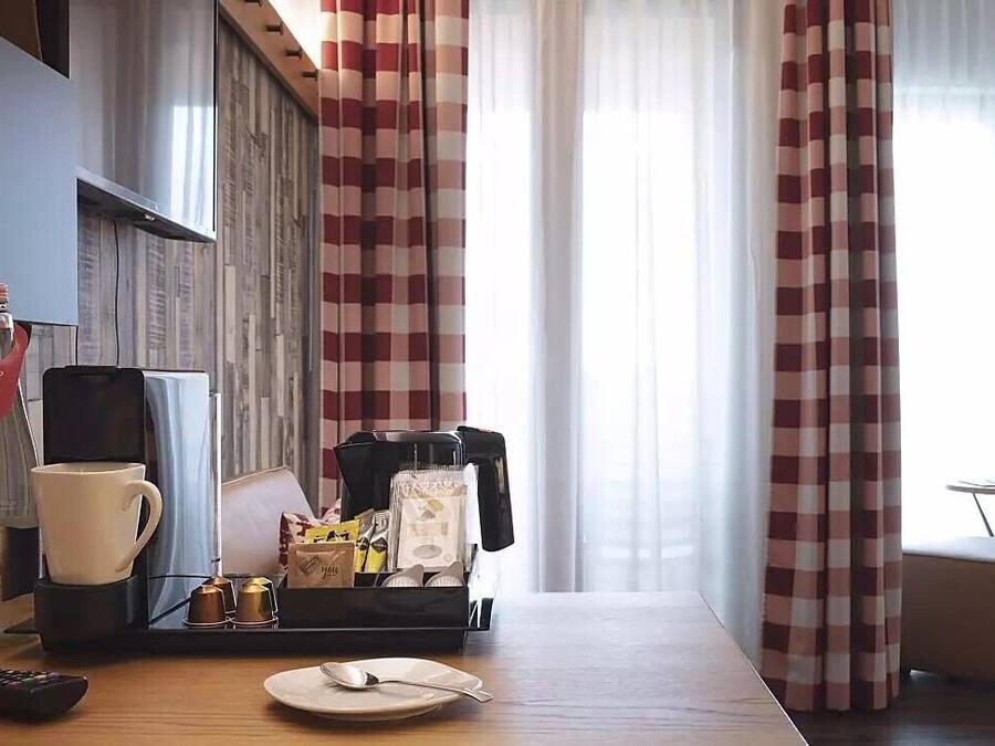 Ein Hotelzimmer mit einem Holztisch, einer Kaffeemaschine, einer Tasse, einem Tablett mit Snacks, einem Bett und Vorhängen.