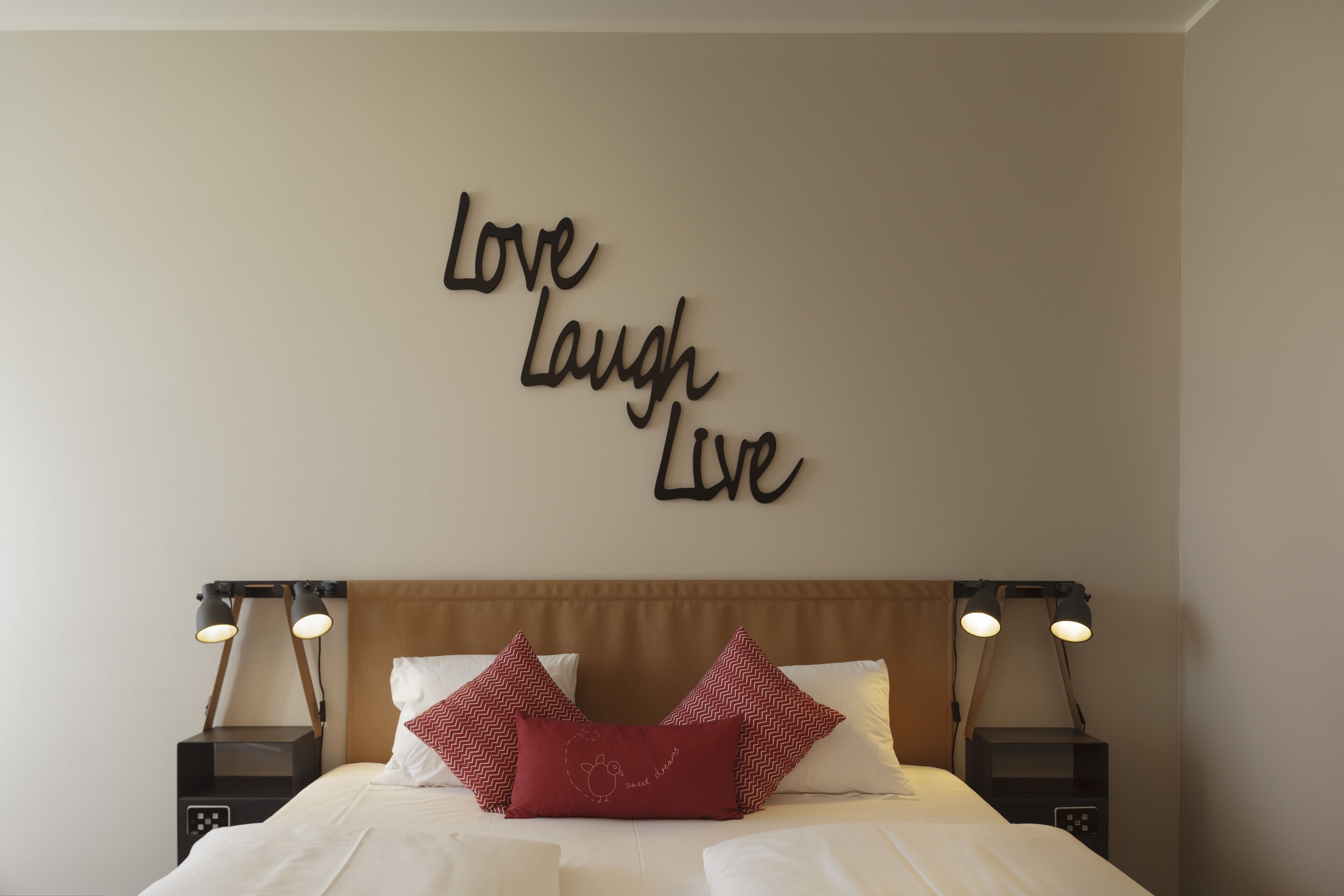 Una cama bien hecha con sábanas blancas y dos almohadas rojas, una pared con una cita inspiradora, y dos lámparas de noche.