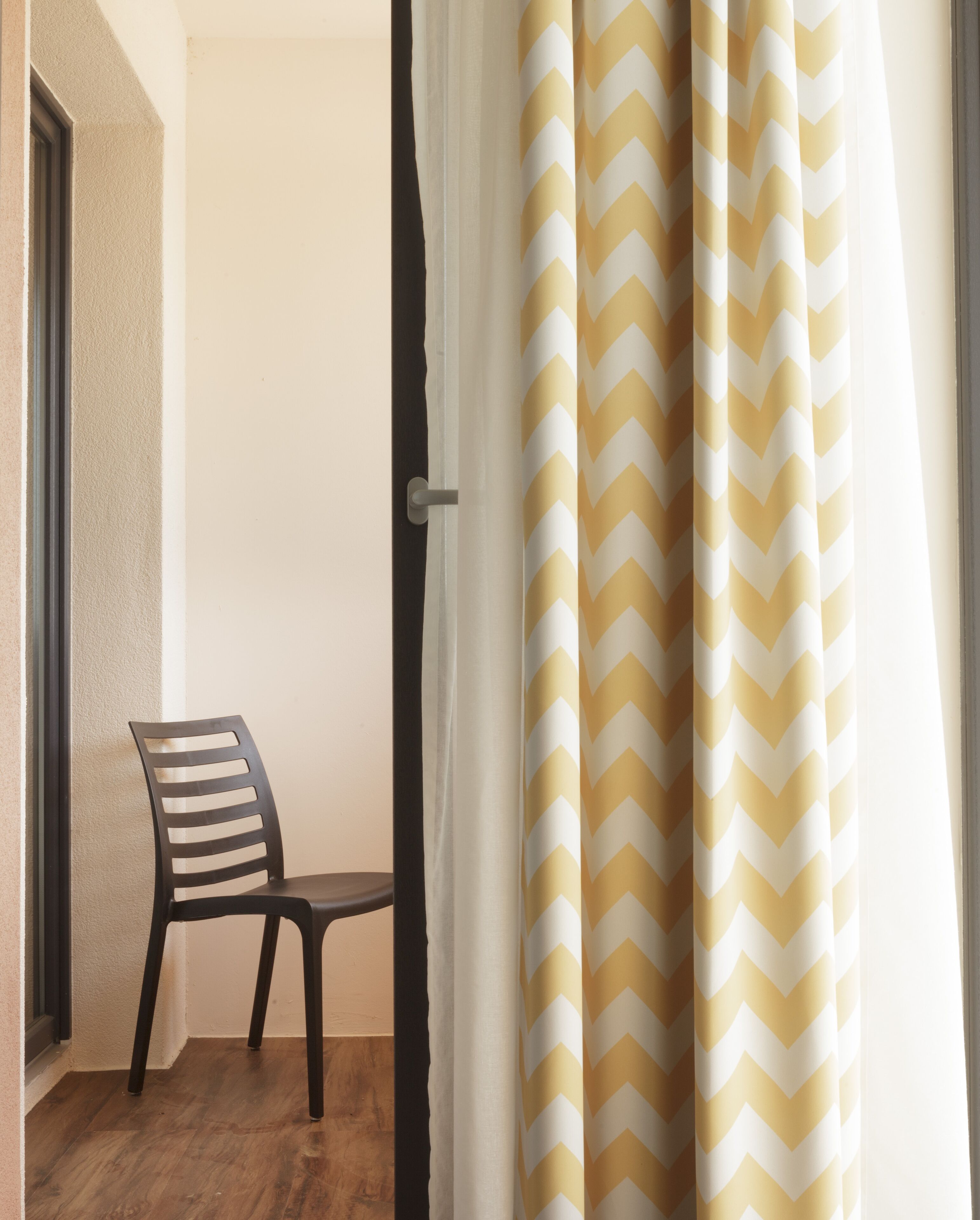 Una habitación con suelo de madera, una silla negra y una ventana con cortina de estampado en zigzag.