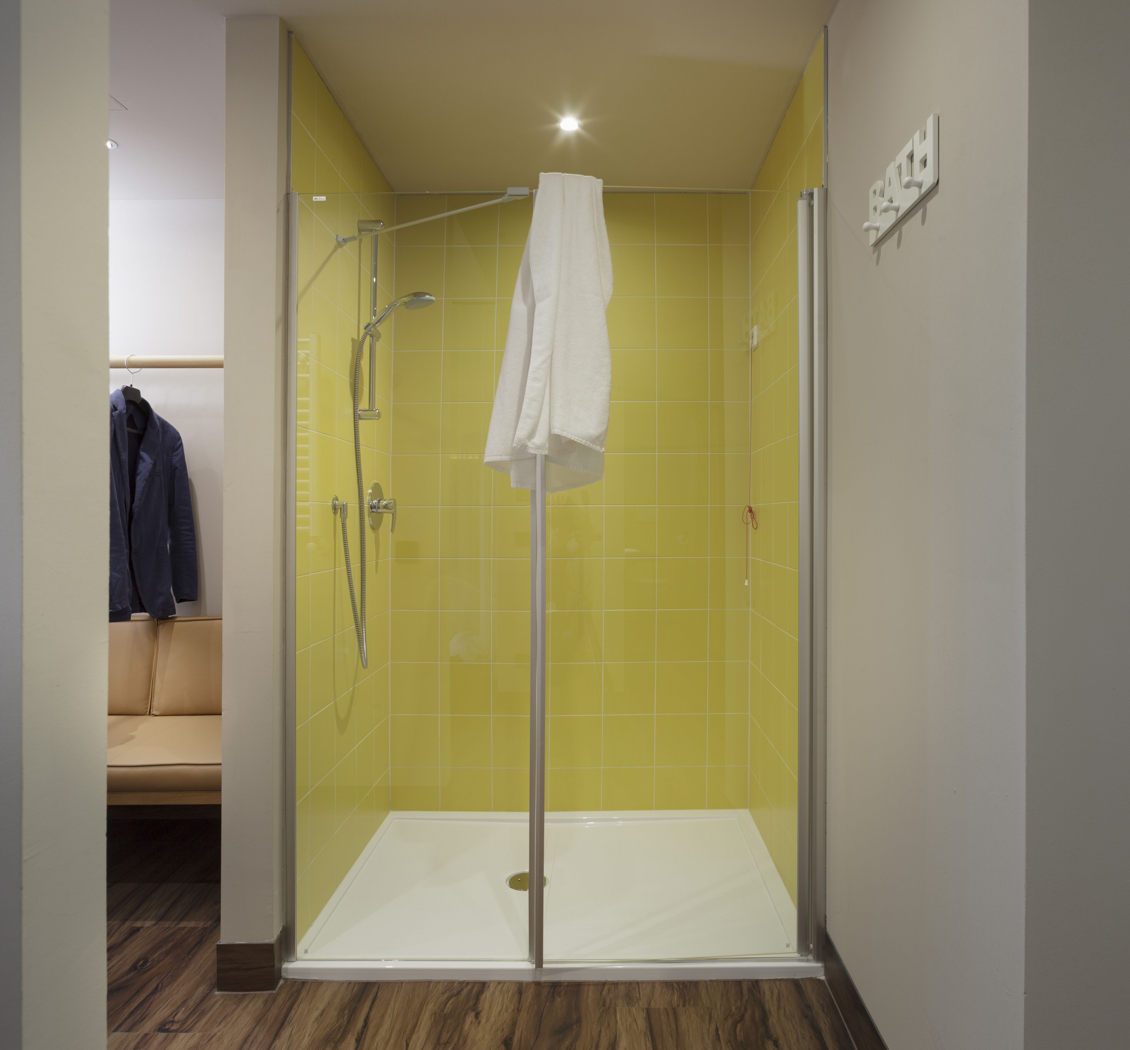 A glass-enclosed shower with a white towel hanging on the door, yellow tiled walls, and a wooden floor.