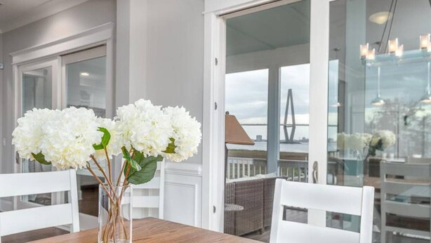 Ein Essbereich mit einem Holztisch, weißen Stühlen, einer Vase mit weißen Blumen und Blick auf eine Brücke durch große Fenster.