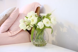 A vase with white flowers on a white table against a wall with a pink pillow.