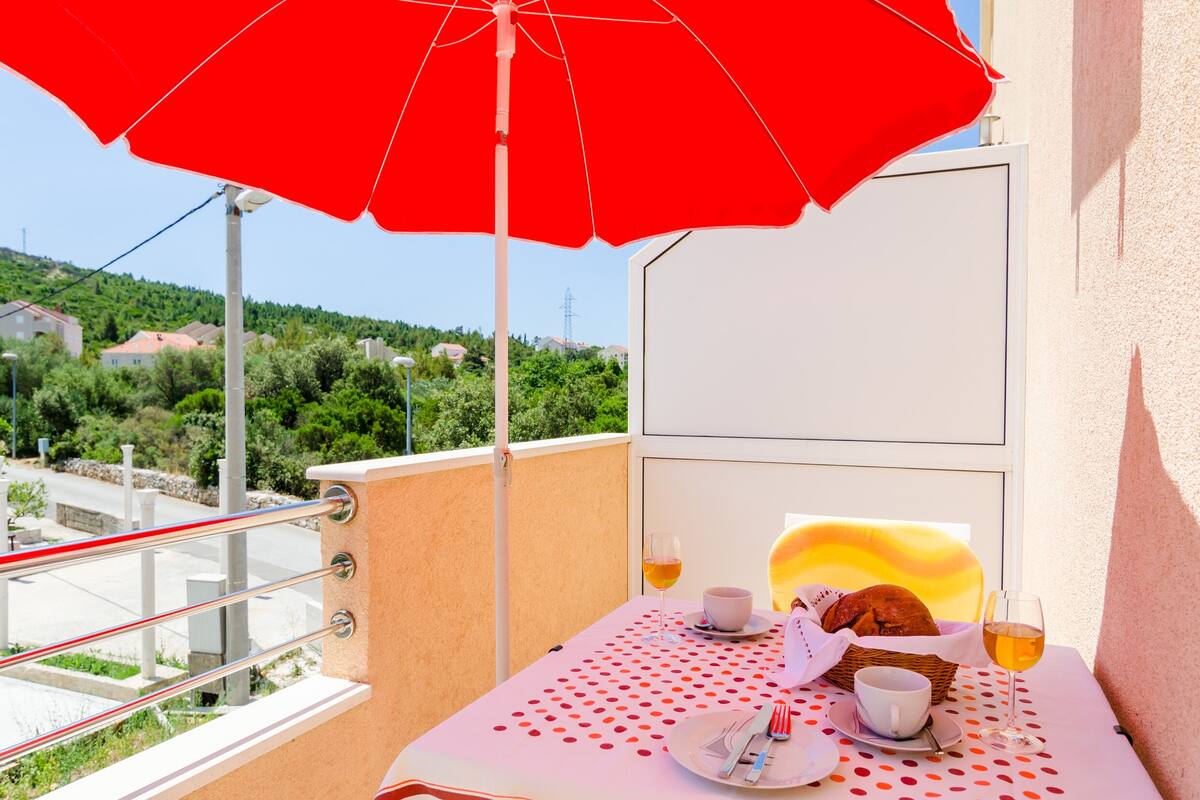 Un balcon avec un parasol rouge, une table recouverte d’une nappe à pois, un panier de pain, une tasse de café et un verre de vin.