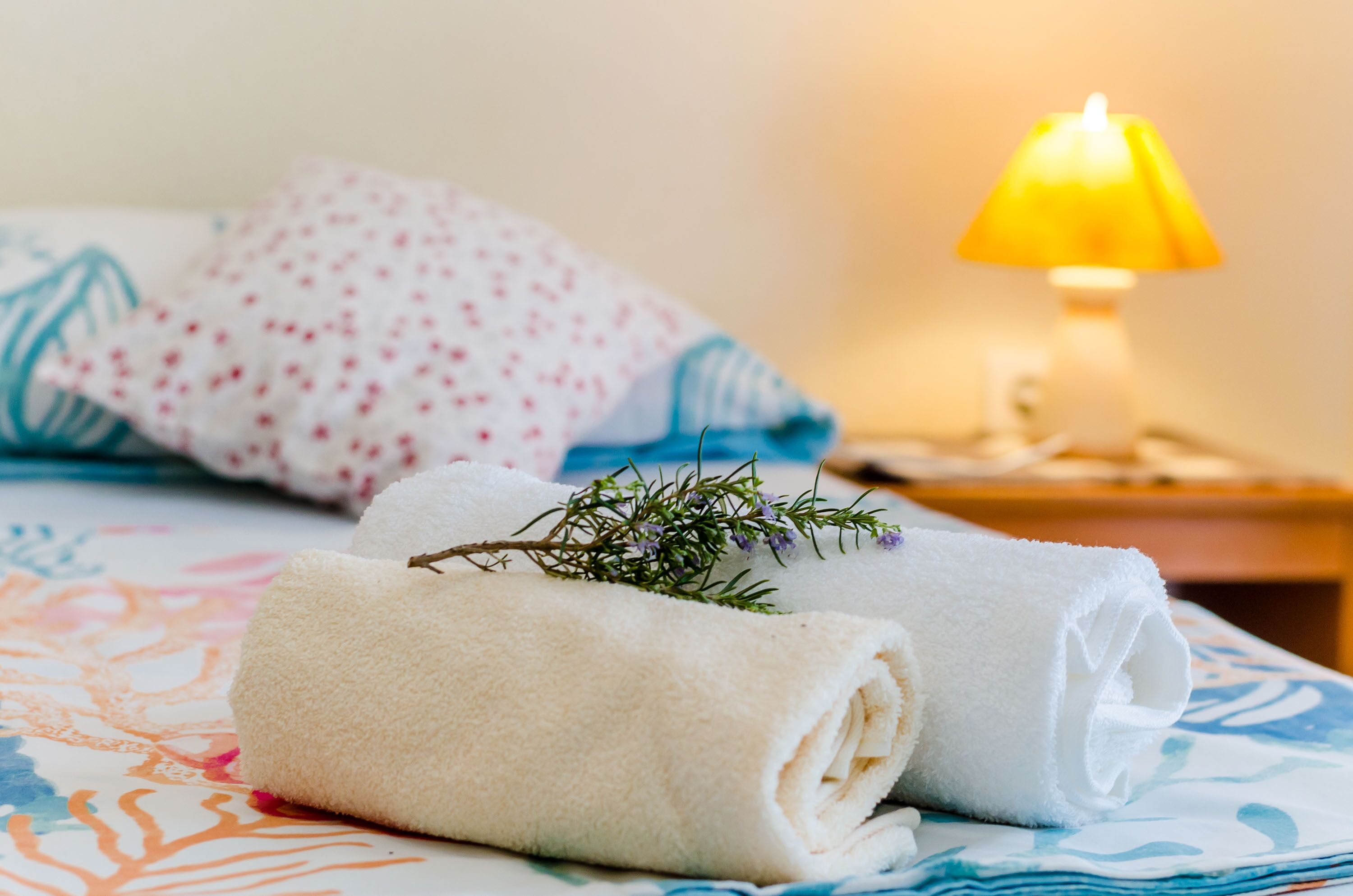 A neatly made bed with rolled white towels and a sprig of rosemary, a patterned pillow, and a bedside lamp with a yellow shade.
