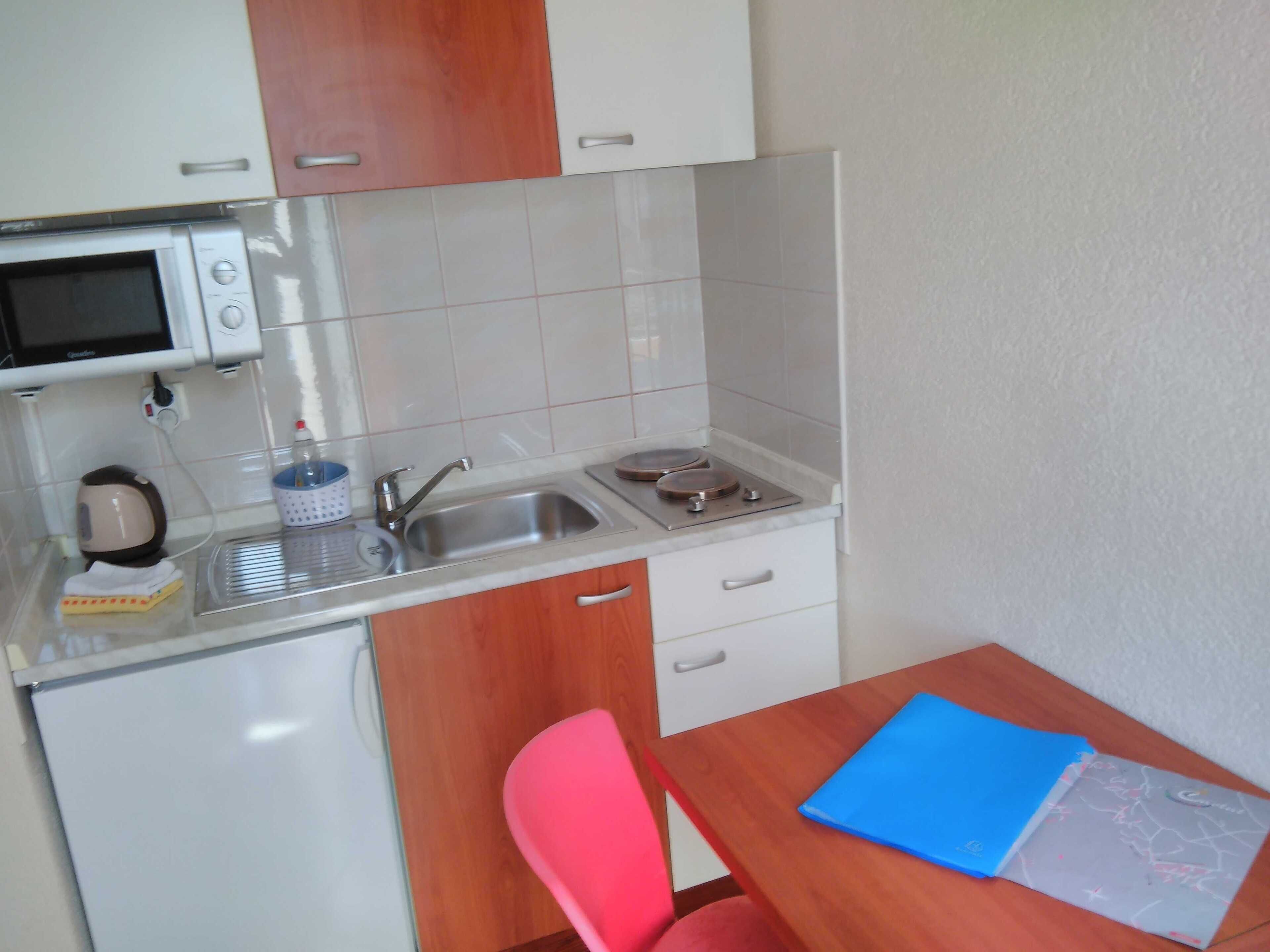 A compact kitchenette with a microwave, sink, and small stove. There is a wooden table with a blue folder and a red chair.