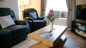 A living room with a sofa, a coffee table with magazines, a TV stand, and a window with curtains. - Eske Holiday Apartment (Donegal Town)