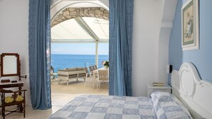 Une chambre avec un grand lit, un bureau et une chaise. On peut admirer la mer à travers une arche ouverte.