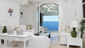 Un salon décoré dans des tons de blanc, avec vue sur la mer, comprenant des canapés blancs, une table basse et une table d’appoint avec une lampe.