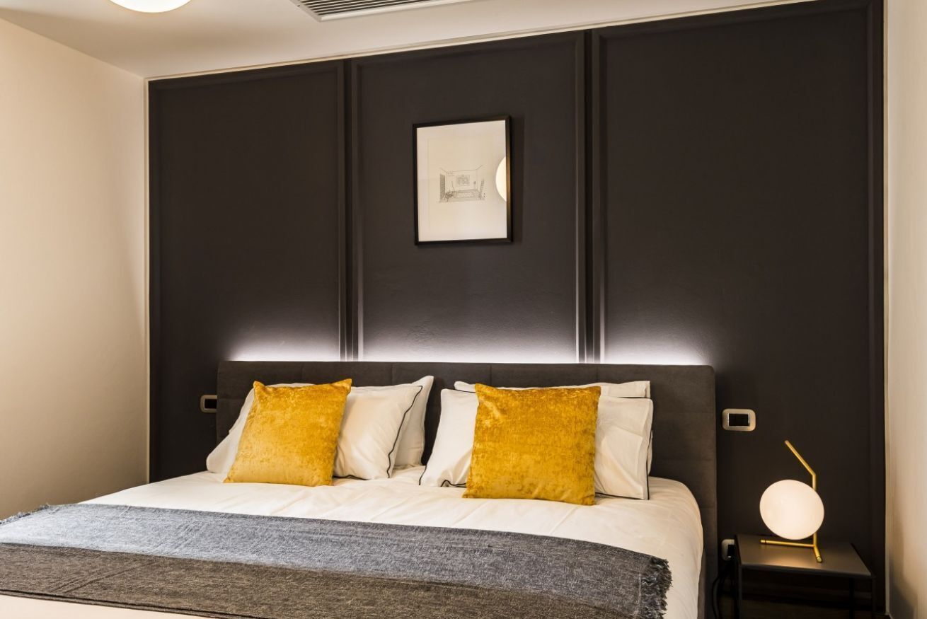 A modern bedroom with a dark headboard, a framed picture, and a bedside lamp.