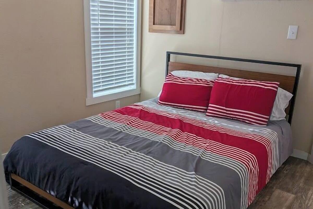 A bedroom with a bed, a window with blinds, an air conditioning unit, and a wooden picture frame on the wall.