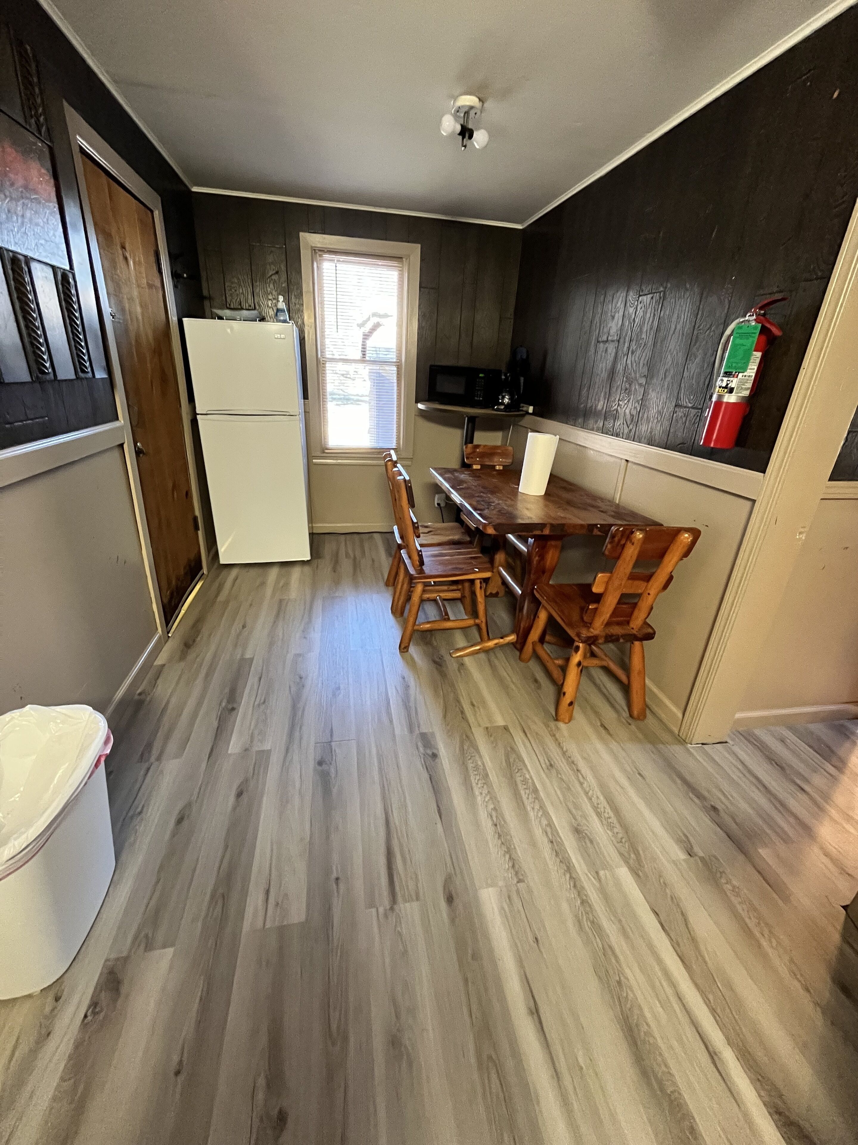A compact kitchen with a wooden dining table and chairs, a white refrigerator, a microwave, and a fire extinguisher on the wall.