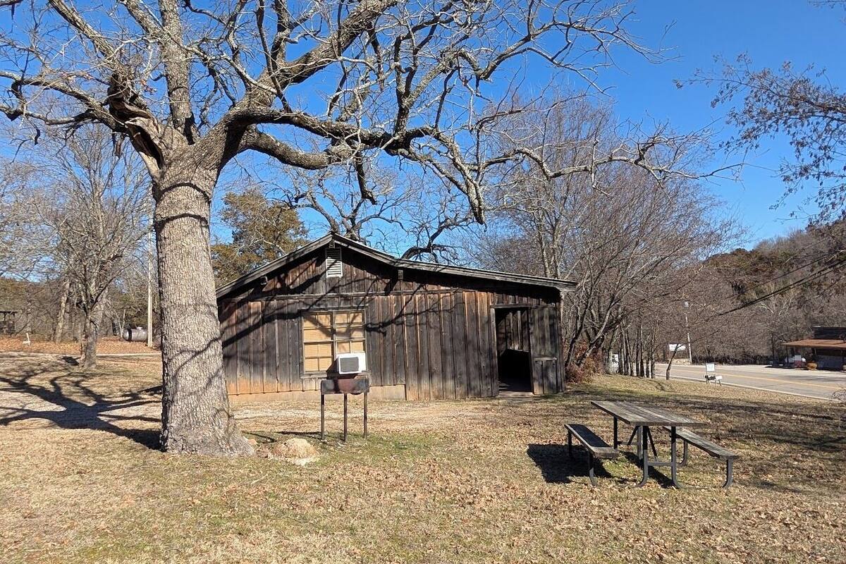 A rustic wooden building with a large tree in front, a picnic table, and a signboard.