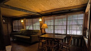 A cozy room with a wooden ceiling, a green sofa, a wooden table with chairs, and large windows with blinds. - Cedarvale Cabins at Turner Falls (Davis)