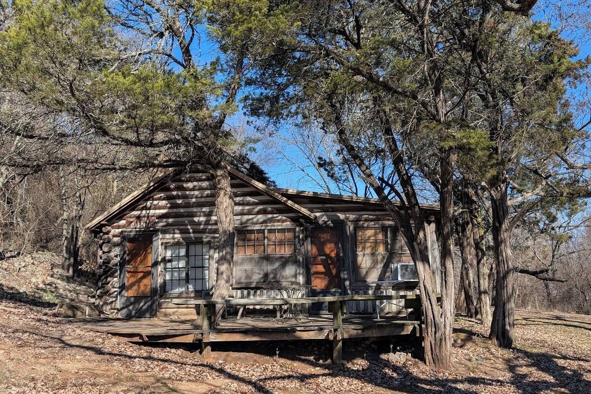 A rustic cabin with a wooden exterior, a front porch, and a large tree in front.