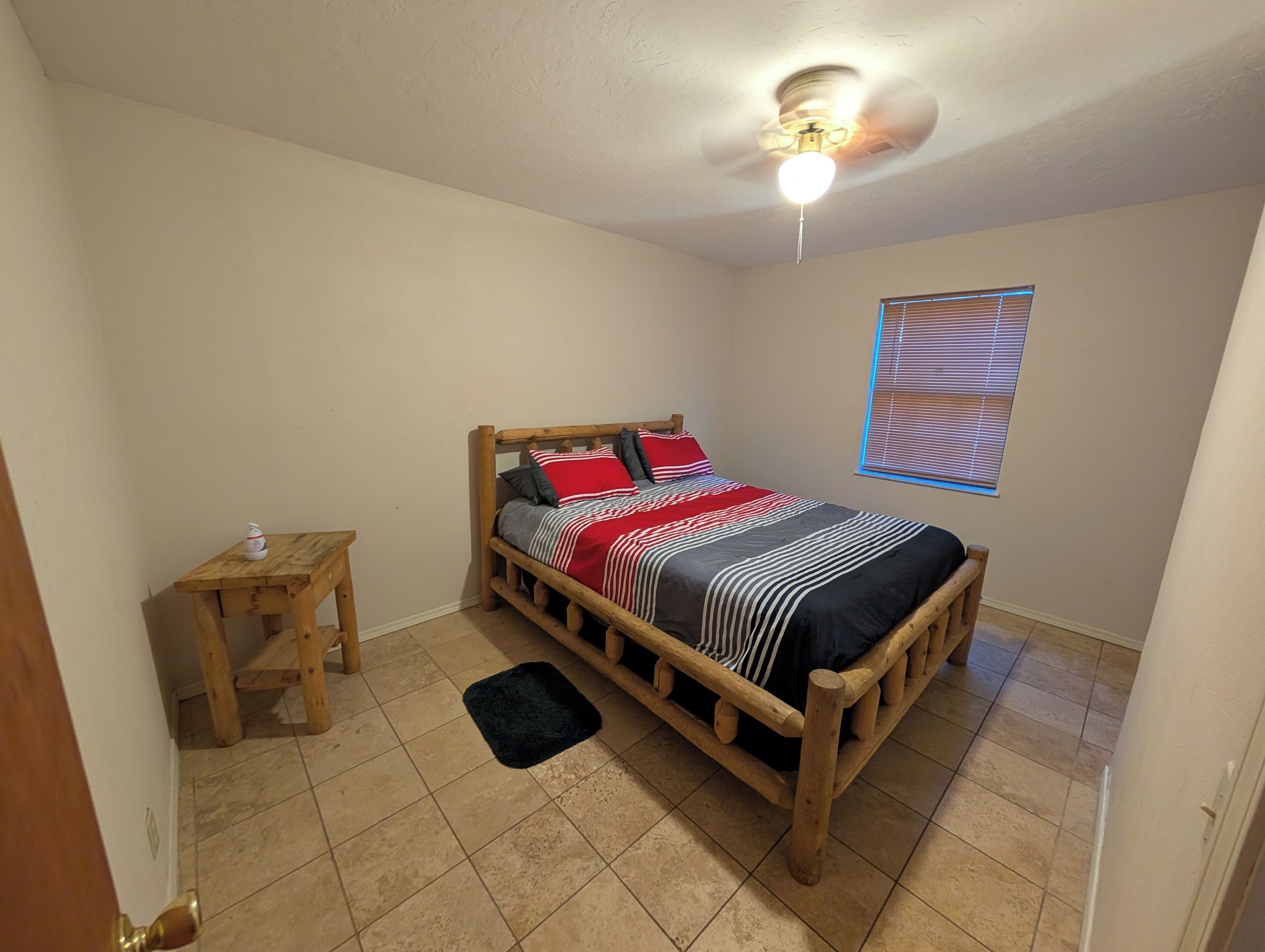 A bedroom with a wooden bed, a small table, a black rug, a ceiling fan, and a window with blinds.