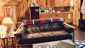 Ein gemütliches Blockhaus-Interieur mit Holzbalkendecke, einer Couch mit gemusterten Decken, einem Esstisch mit Lampe und einer Küchenzeile inklusive Mikrowelle und Kühlschrank.