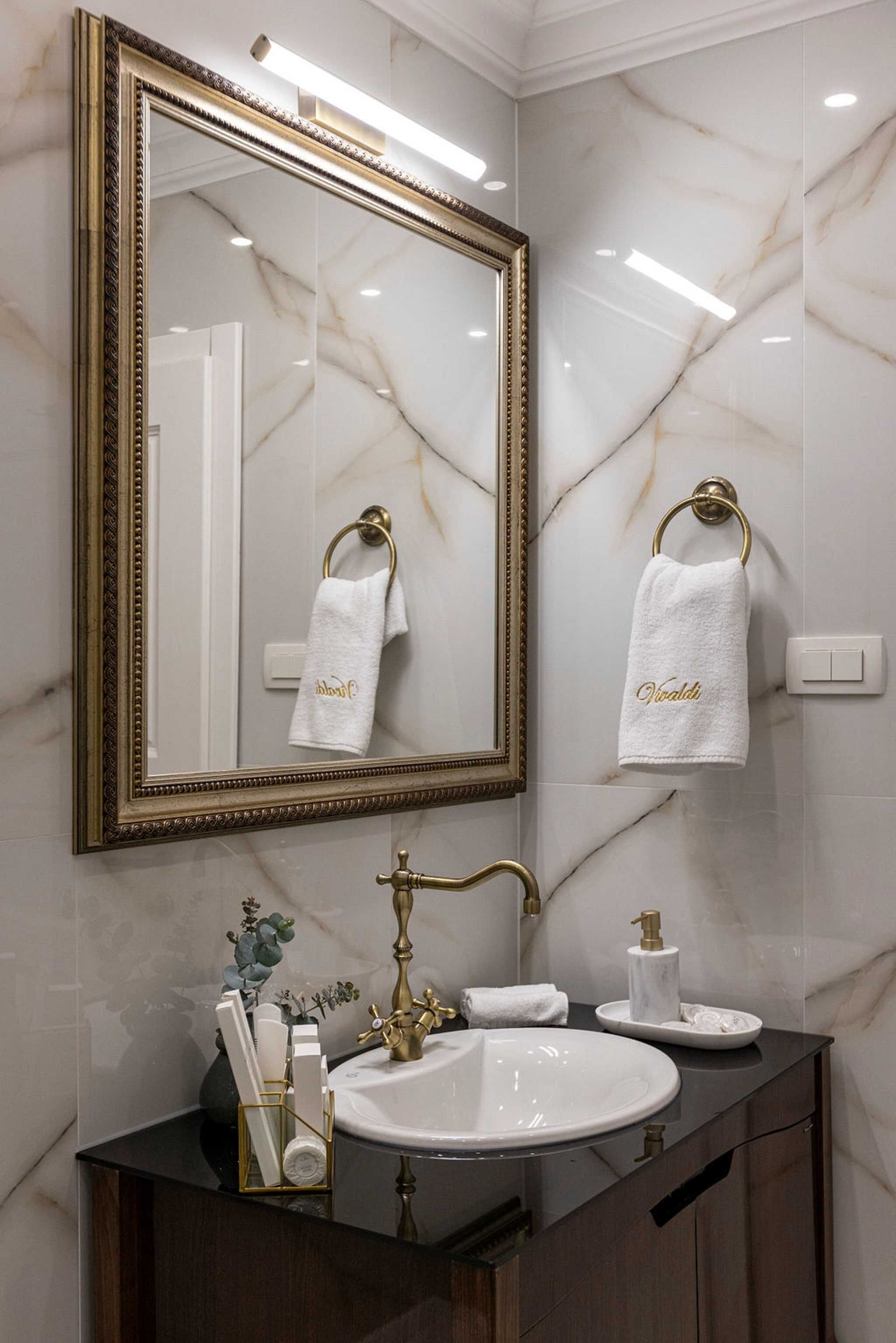 A bathroom with a marble wall, a gold-framed mirror, a white sink, and gold faucets.
