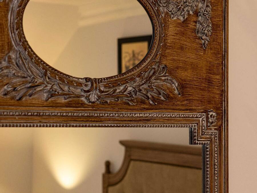 Een spiegel met houten frame en gedetailleerd snijwerk, een bed met een gestoffeerde hoofdbord en een bedlampje.