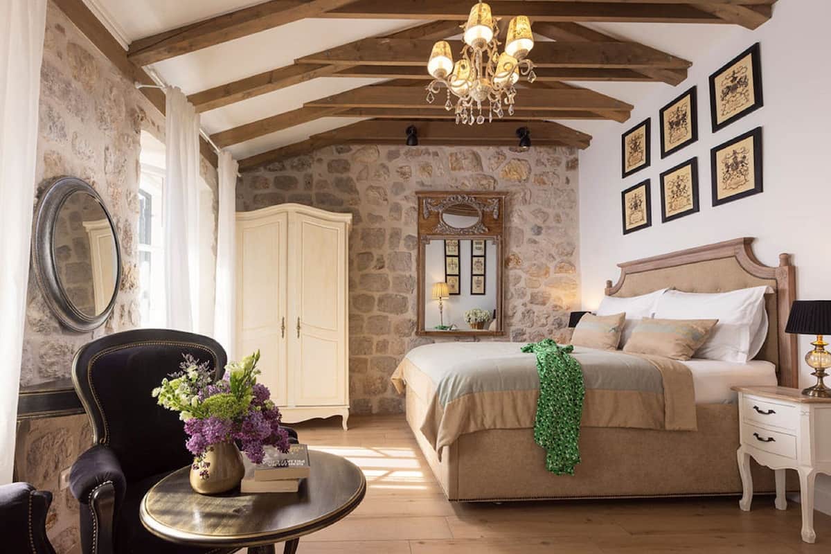 A bedroom with a stone wall, a bed with a green throw, a chandelier, and framed pictures on the wall.