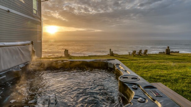 A hot tub with a wooden deck, overlooking a grassy area and the ocean at sunset. - Oceanfront Whale Watching Retreat (Depoe Bay)