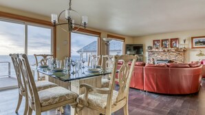 A dining area with a glass table and chairs, a kitchen with a stone fireplace, and a view of the ocean. - Oceanfront Whale Watching Retreat (Depoe Bay)