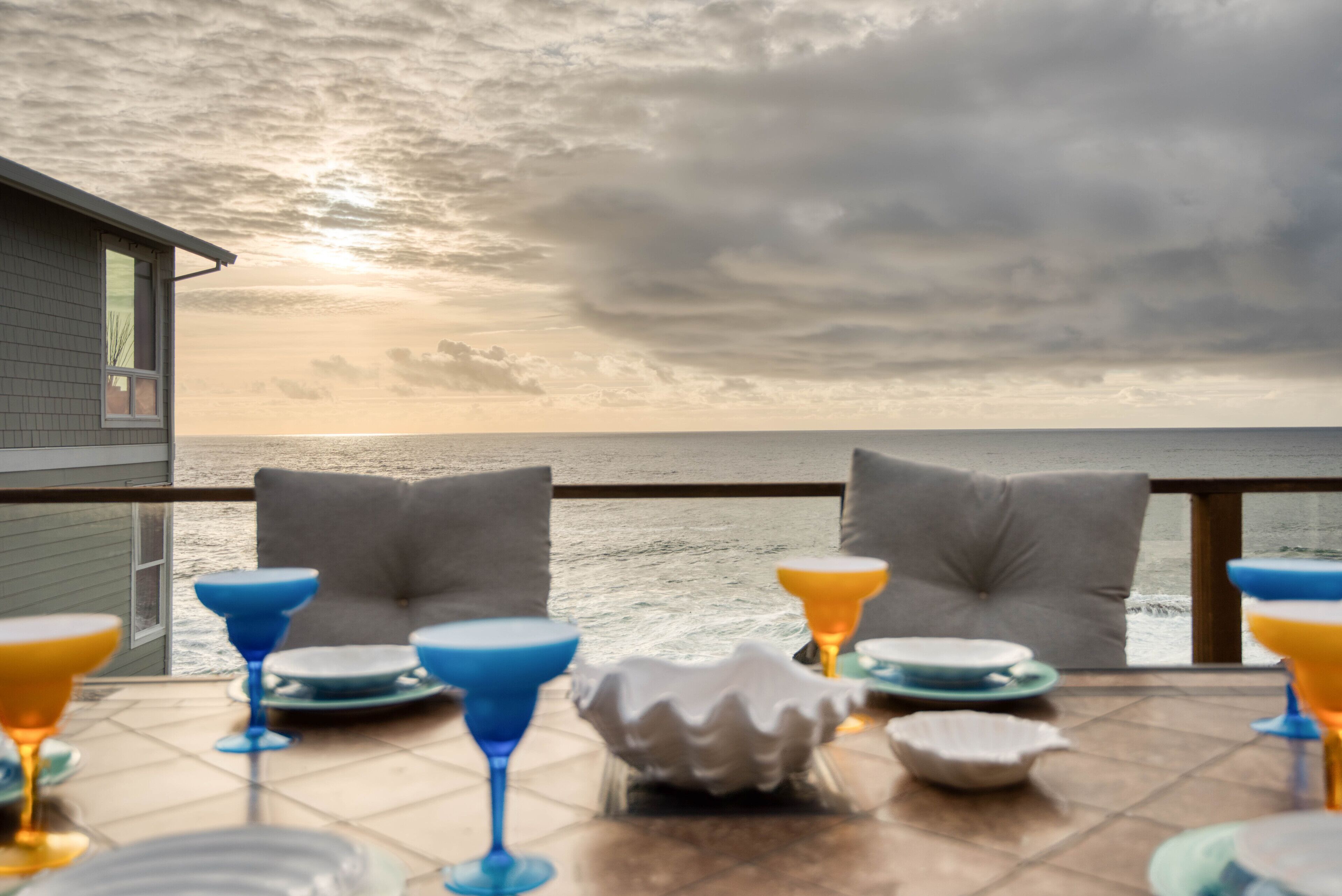 Une table dressée avec des verres à cocktail bleus et oranges, des assiettes et un saladier blanc, donnant sur une étendue d’eau sous un ciel nuageux.