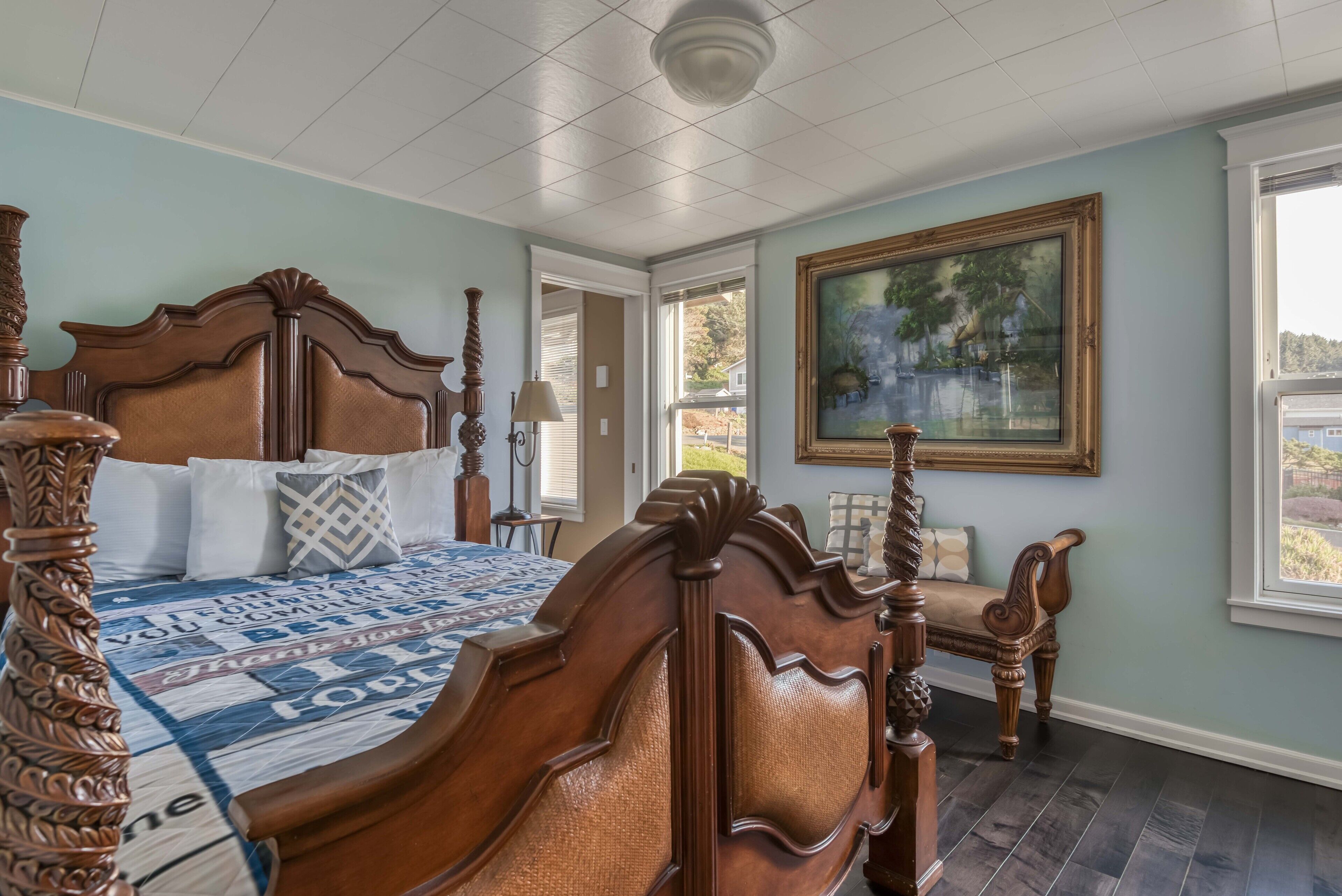 A bedroom with a large bed, a wooden chair, a painting on the wall, and a view through the window.