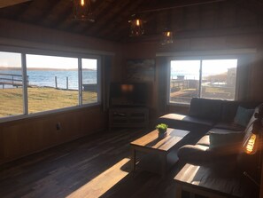 Una sala de estar acogedora con suelo de madera, un sofá, una mesa de centro y un televisor. Amplias ventanas ofrecen vistas al exterior.