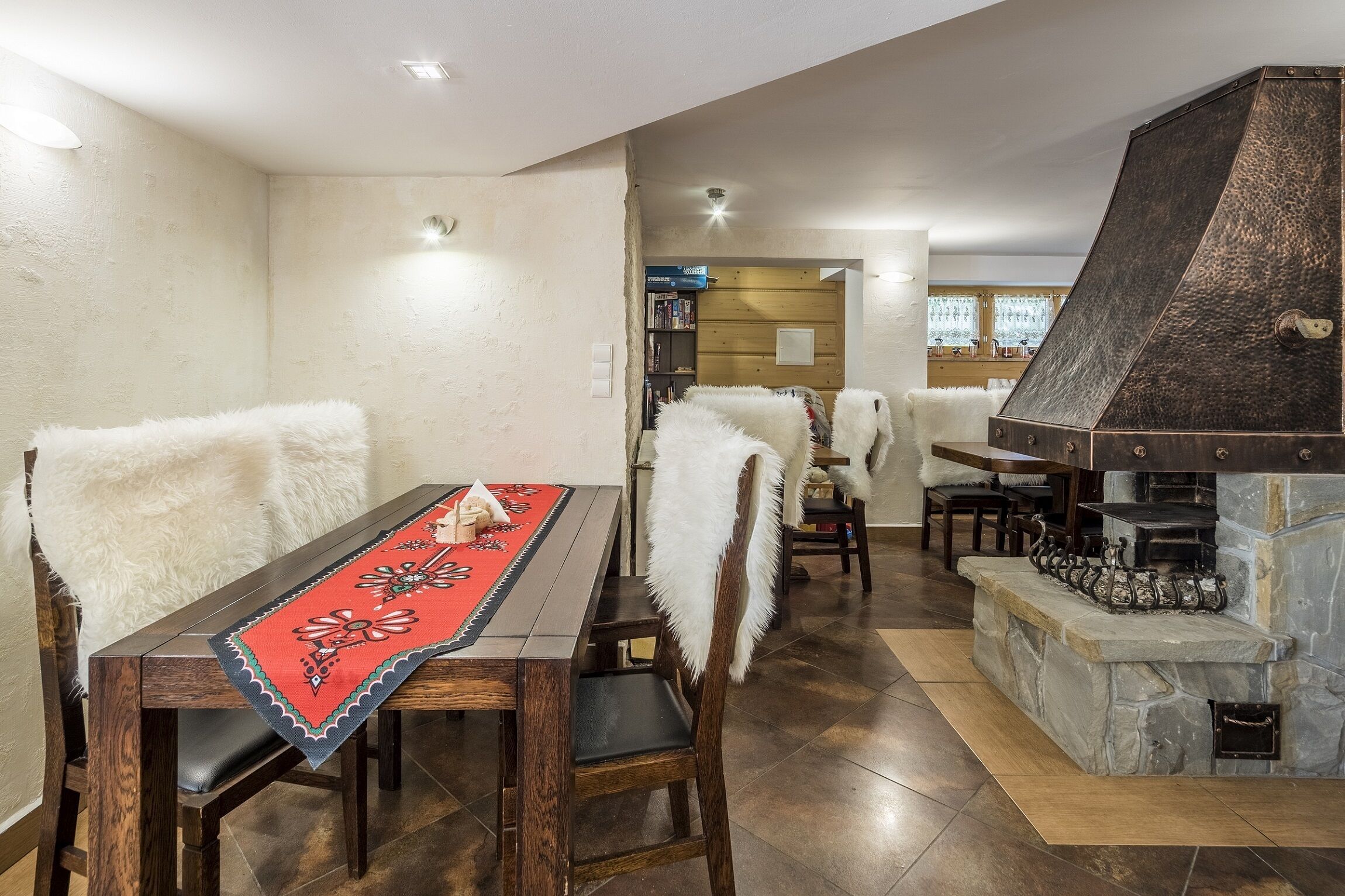 A dining area with a wooden table and chairs, a red table runner, and a stone fireplace.