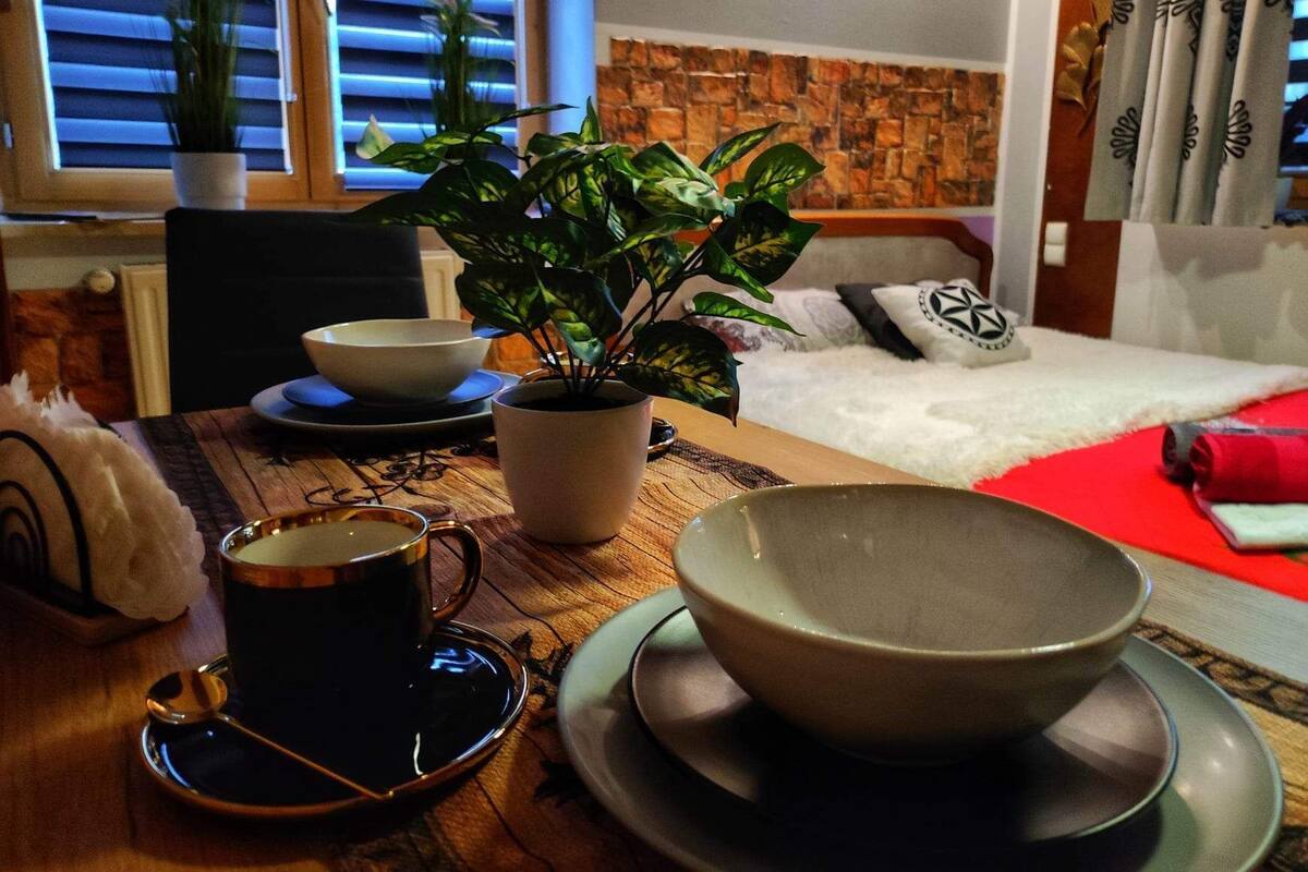 Ein gemütliches Zimmer mit einem Holztisch, einer Topfpflanze, einer Tasse Kaffee und einem Bett mit roter Decke.