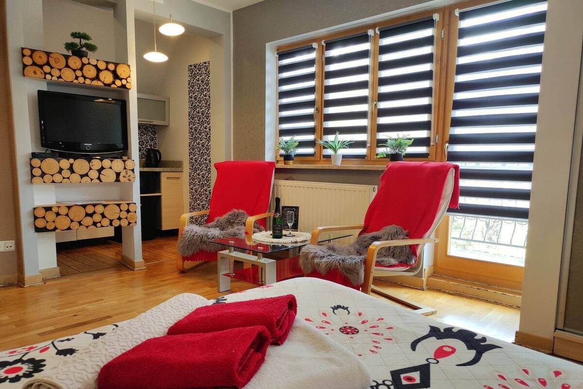 Ein modernes Hotelzimmer mit einem Bett, zwei roten Sessel, einem kleinen Tisch mit Flasche und Glas, einem Fernseher auf einem Regal und einem Fenster mit Jalousien.