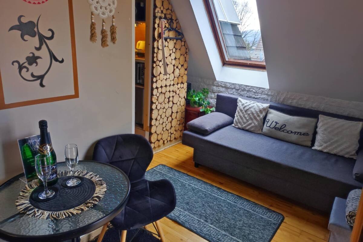 Ein gemütliches Zimmer mit einem grauen Sofa, einem kleinen runden Tisch mit zwei Weingläsern und einer Flasche, sowie einer Wand mit einem Traumfänger.