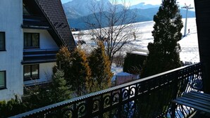 Un immeuble résidentiel doté d’un toit en pente, d’un balcon orné de rampes en métal ouvragé et offrant une vue sur un paysage enneigé avec des montagnes en arrière-plan.