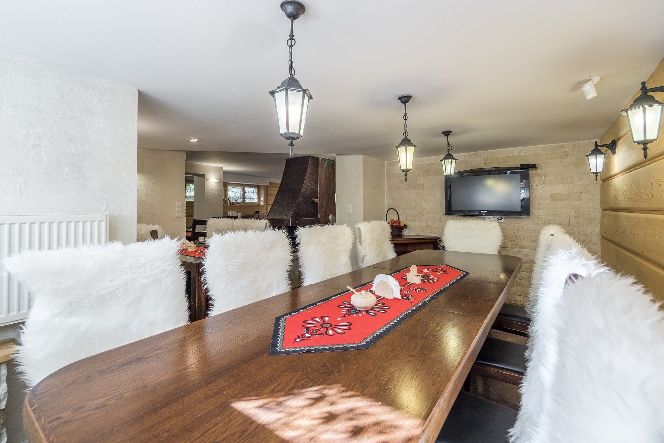 A dining area with a long wooden table, white furry chairs, and a red placemat. There are hanging lantern-style lights and a flat-screen TV mounted on the wall.