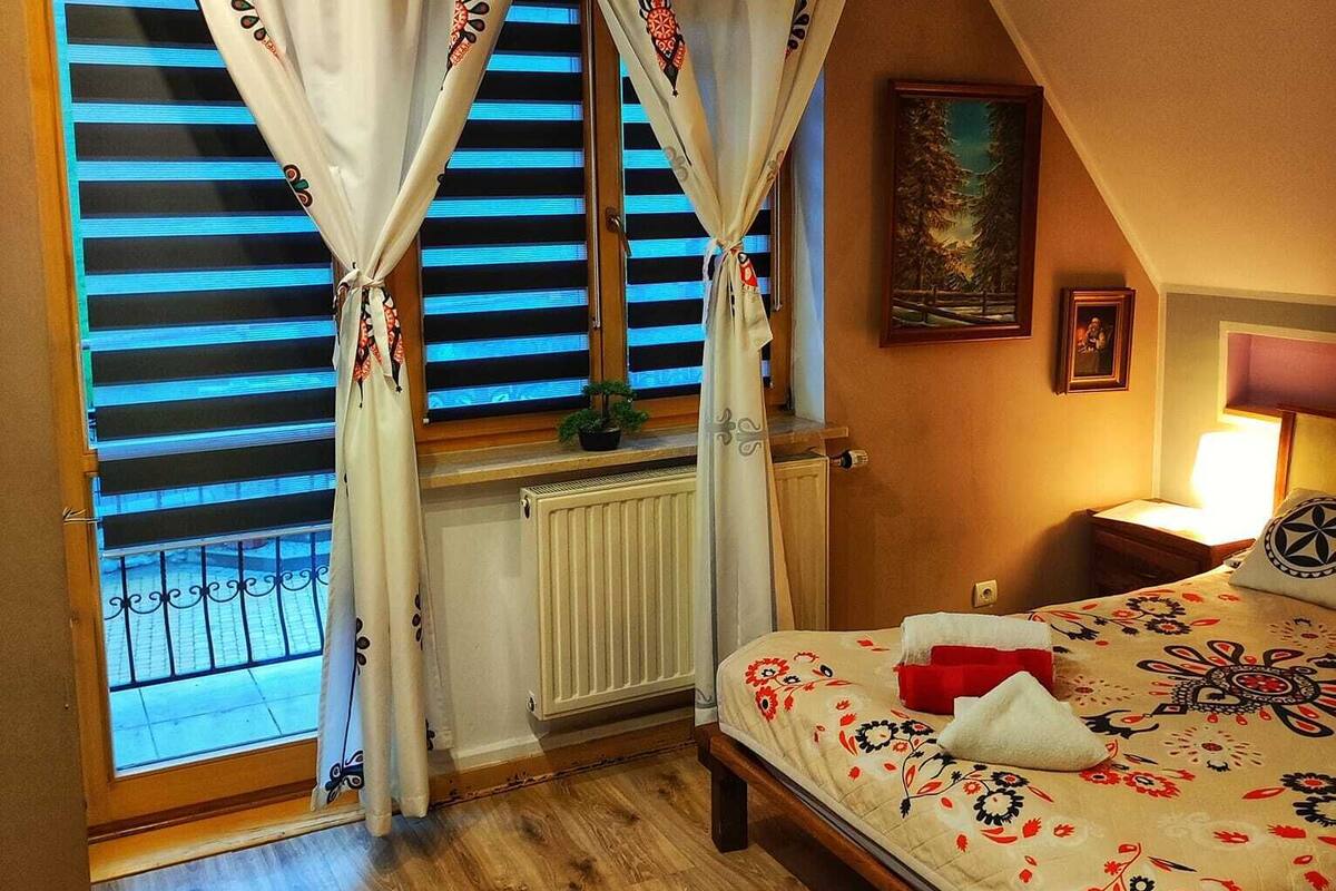 Ein Schlafzimmer mit Bett, einem Fenster mit Vorhängen, einem Heizkörper, einem Bild an der Wand und einem Nachttisch mit Lampe.