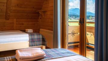 A wooden cabin room with two beds, a window with a view of mountains, and a wooden door.