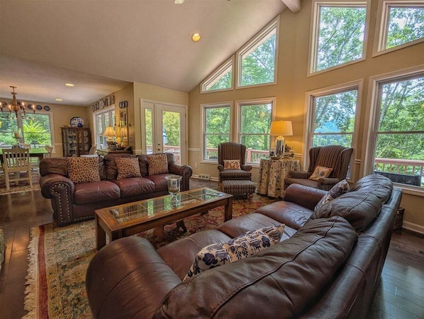 A spacious living room with leather sofas, a glass coffee table, and large windows offering a view of greenery.