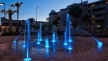 Escena nocturna con varias fuentes iluminadas con luces azules, rodeadas de palmeras y una zona pavimentada con ladrillos.