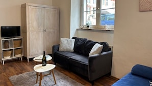 Een woonkamer met een donker leren bank, een witte kast, een televisie en een raam met uitzicht op gebouwen.