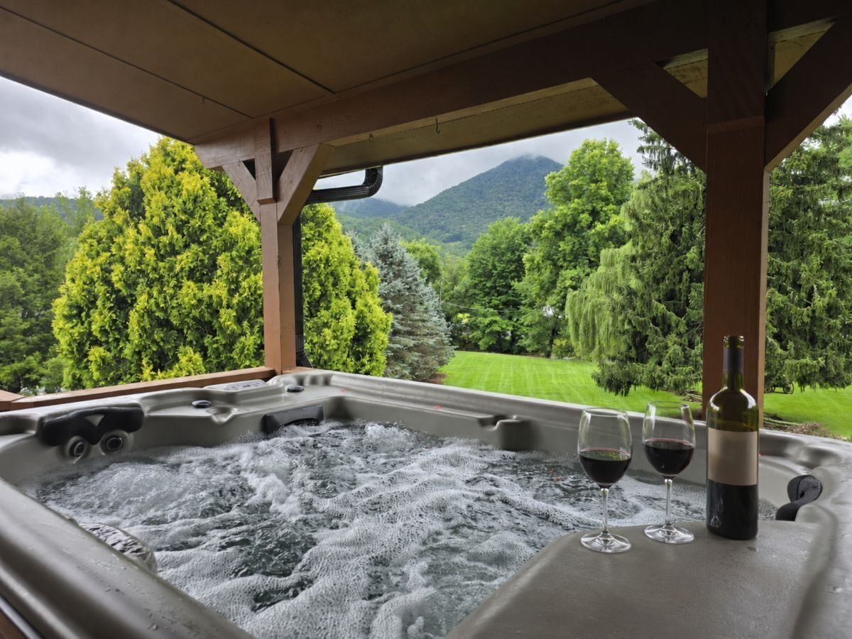 A hot tub with two glasses of red wine and a bottle, set against a backdrop of lush greenery and mountains.