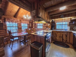 Interior rústico de una cabaña con una mesa de comedor de madera, sillas, una cocina con estufa y un lavamanos.