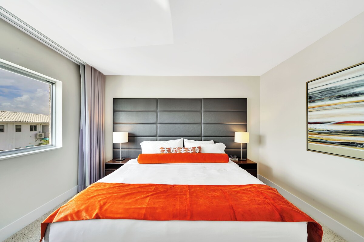 Une chambre d’hôtel avec un grand lit, une tête de lit, deux tables de chevet avec des lampes, une fenêtre avec des rideaux et un tableau au mur.
