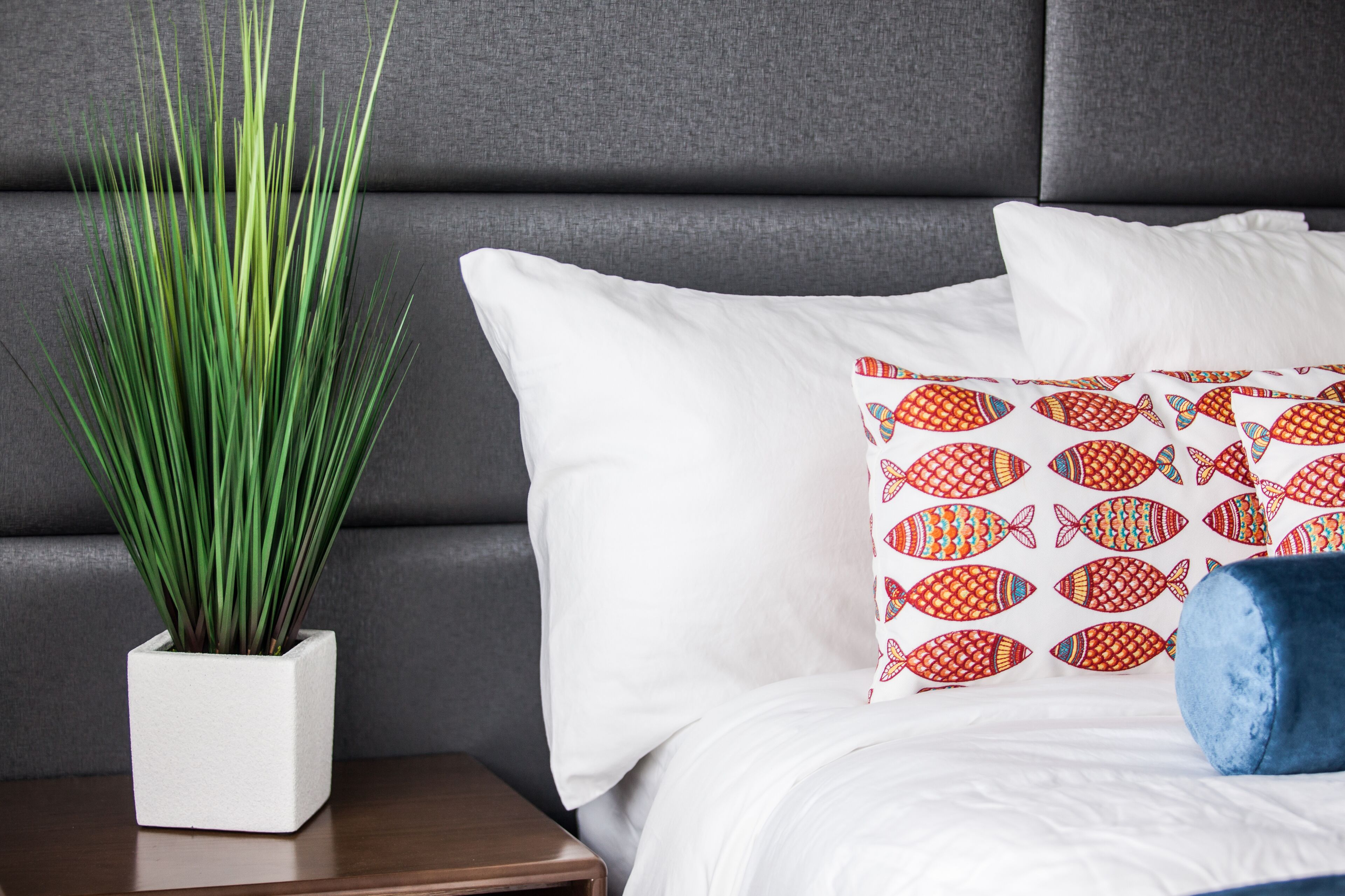 A neatly arranged bedroom with a grey upholstered headboard, white pillows, a decorative fish-patterned pillow, a blue cushion, and a potted plant on a wooden nightstand.