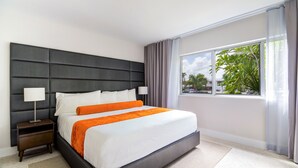 A modern hotel room with a large bed, a nightstand, a window with a view of greenery, and a grey headboard. - The Elmar Boutique Hotel (Lauderdale-by-the-Sea)
