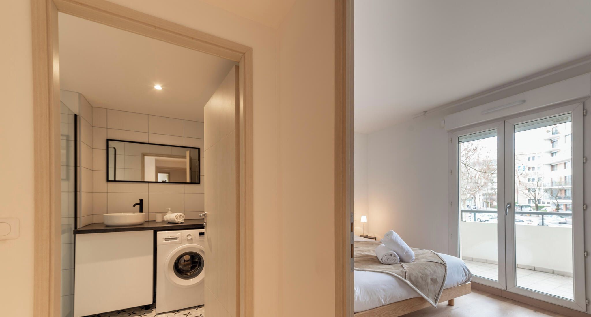 Un apartamento moderno con baño, lavadora, cama y balcón con vista a edificios.