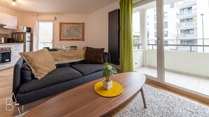 Un saló modern amb un sofà negre, una taula de centre de fusta i una planta en test. Hi ha una zona de cuina amb microones i tetera, i un balcó amb vista a edificis.