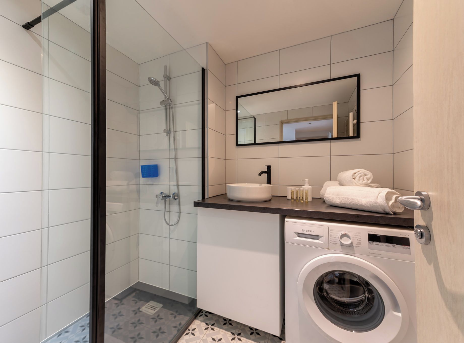 Een moderne badkamer met een douchecabine, een Bosch wasmachine, een wastafel met spiegel erboven en een handdoekhouder.