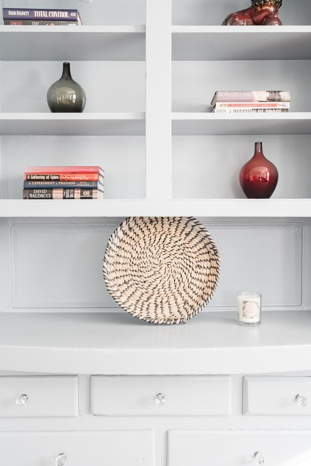A white shelf with books, a red vase, a woven basket, and a candle.